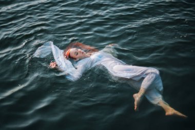 Güzel kızıl saçlı kız suda yüzüyor. Kayalık sahil. Bir model beyaz bir antika elbise zümrüt suda. Ortaçağ görüntüsü. Fantezi