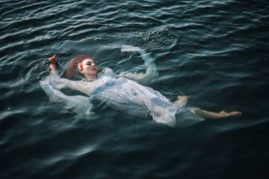 Güzel kızıl saçlı kız suda yüzüyor. Kayalık sahil. Bir model beyaz bir antika elbise zümrüt suda. Ortaçağ görüntüsü. Fantezi