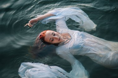 Güzel kızıl saçlı kız suda yüzüyor. Kayalık sahil. Bir model beyaz bir antika elbise zümrüt suda. Ortaçağ görüntüsü. Fantezi
