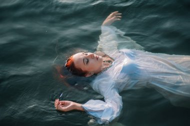 Güzel kızıl saçlı kız suda yüzüyor. Kayalık sahil. Bir model beyaz bir antika elbise zümrüt suda. Ortaçağ görüntüsü. Fantezi