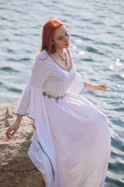 Güzel kızıl saçlı kız suda yüzüyor. Kayalık sahil. Bir model beyaz bir antika elbise zümrüt suda. Ortaçağ görüntüsü. Fantezi