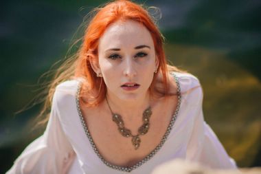 Güzel kızıl saçlı kız suda yüzüyor. Kayalık sahil. Bir model beyaz bir antika elbise zümrüt suda. Ortaçağ görüntüsü. Fantezi