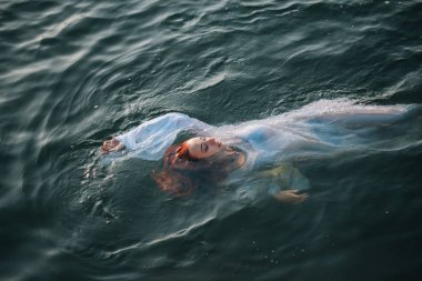 Güzel kızıl saçlı kız suda yüzüyor. Kayalık sahil. Bir model beyaz bir antika elbise zümrüt suda. Ortaçağ görüntüsü. Fantezi