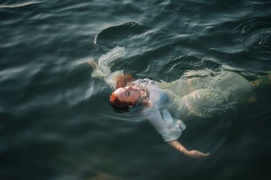 Güzel kızıl saçlı kız suda yüzüyor. Kayalık sahil. Bir model beyaz bir antika elbise zümrüt suda. Ortaçağ görüntüsü. Fantezi