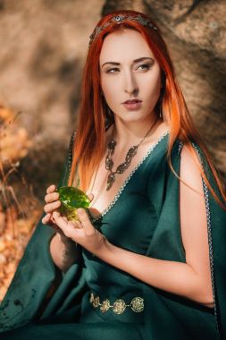 Turkuaz elbise ve bir diadem başında güzel bir kızıl saçlı kız sonbahar ormanda yürüyor. Bir elf görüntüde. Bu dağlarda büyük kayalar. Güneş parlamayı. Fantezi