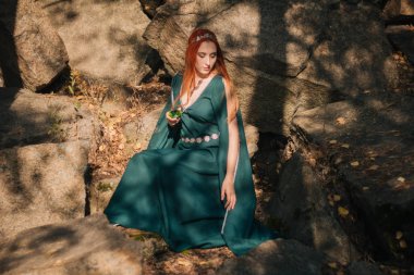 Turkuaz elbise ve bir diadem başında güzel bir kızıl saçlı kız sonbahar ormanda yürüyor. Bir elf görüntüde. Bu dağlarda büyük kayalar. Güneş parlamayı. Fantezi