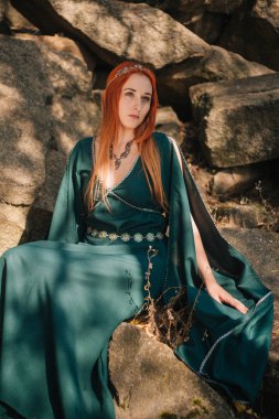 Turkuaz elbise ve bir diadem başında güzel bir kızıl saçlı kız sonbahar ormanda yürüyor. Bir elf görüntüde. Bu dağlarda büyük kayalar. Güneş parlamayı. Fantezi