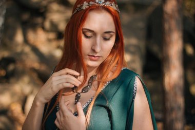 Turkuaz elbise ve bir diadem başında güzel bir kızıl saçlı kız sonbahar ormanda yürüyor. Bir elf görüntüde. Bu dağlarda büyük kayalar. Güneş parlamayı. Fantezi