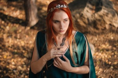 Turkuaz elbise ve bir diadem başında güzel bir kızıl saçlı kız sonbahar ormanda yürüyor. Bir elf görüntüde. Bu dağlarda büyük kayalar. Güneş parlamayı. Fantezi