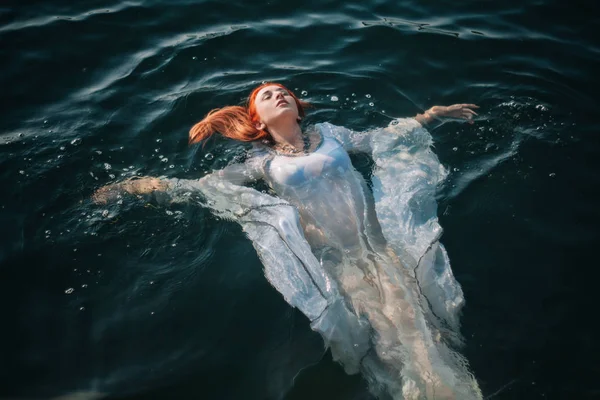 Güzel kızıl saçlı kız suda yüzüyor. Kayalık sahil. Bir model beyaz bir antika elbise zümrüt suda. Ortaçağ görüntüsü. Fantezi