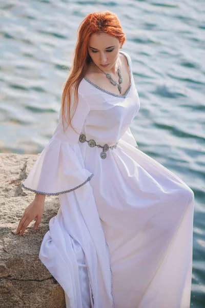 Beautiful red-haired girl is floating in the water. Rocky coast. A model in emerald water in a white antique dress. Medieval image. Fantasy