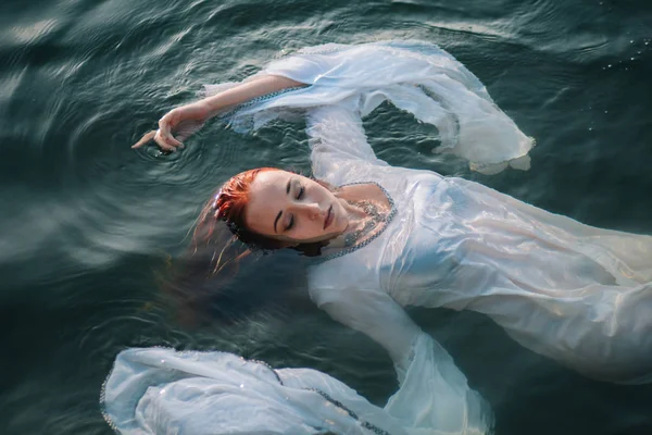 Güzel kızıl saçlı kız suda yüzüyor. Kayalık sahil. Bir model beyaz bir antika elbise zümrüt suda. Ortaçağ görüntüsü. Fantezi