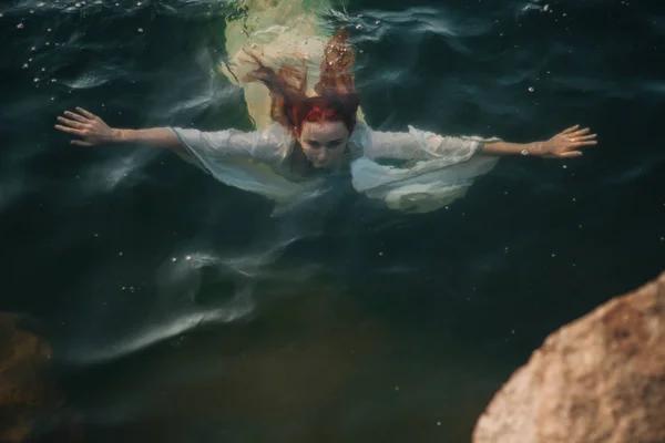 Beautiful red-haired girl is floating in the water. Rocky coast. A model in emerald water in a white antique dress. Medieval image. Fantasy