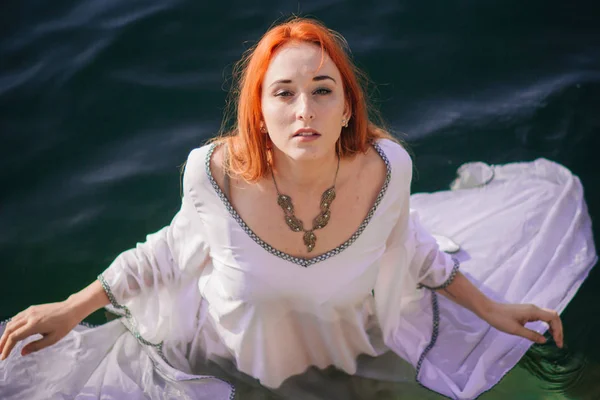 Beautiful red-haired girl is floating in the water. Rocky coast. A model in emerald water in a white antique dress. Medieval image. Fantasy