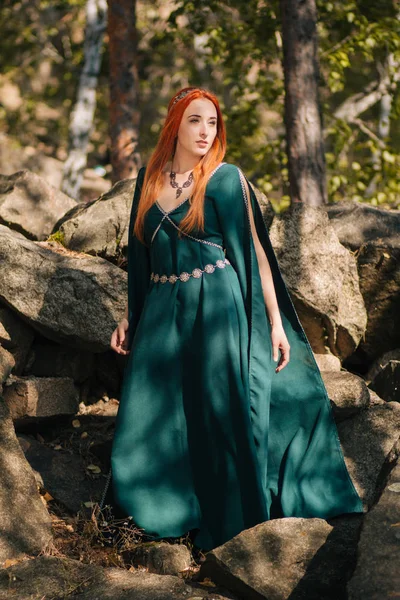 A beautiful red-haired girl in a turquoise dress and a diadem on her head is walking in the autumn forest. In the image of an elf. It is on large rocks in the mountains. Solar glare. Fantasy