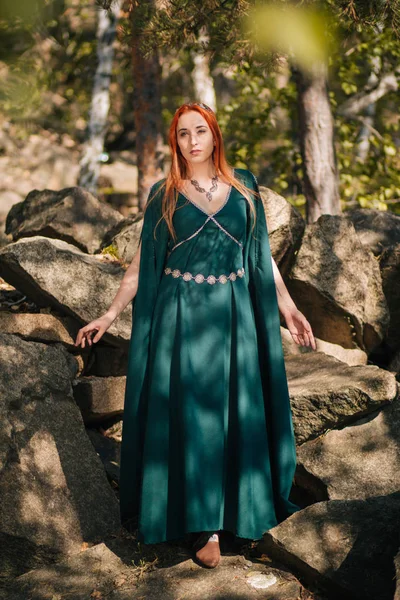 ターコイズ ブルーのドレスと彼女の頭の上の王冠の美しい赤い髪の少女は 秋の森で歩いています エルフのイメージ 山の中の大きな岩です 太陽のまぶしさ ファンタジー — ストック写真