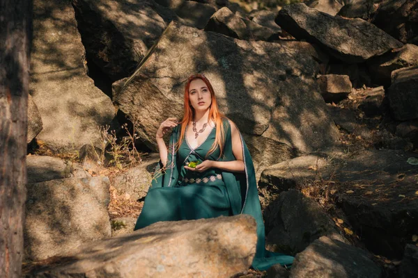 ターコイズ ブルーのドレスと彼女の頭の上の王冠の美しい赤い髪の少女は 秋の森で歩いています エルフのイメージ 山の中の大きな岩です 太陽のまぶしさ ファンタジー — ストック写真