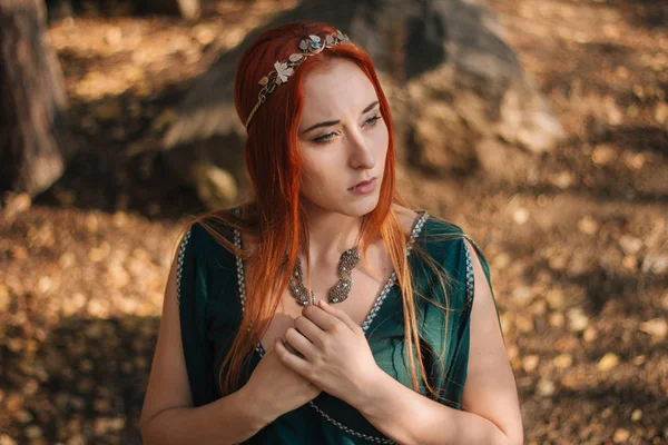 A beautiful red-haired girl in a turquoise dress and a diadem on her head is walking in the autumn forest. In the image of an elf. It is on large rocks in the mountains. Solar glare. Fantasy