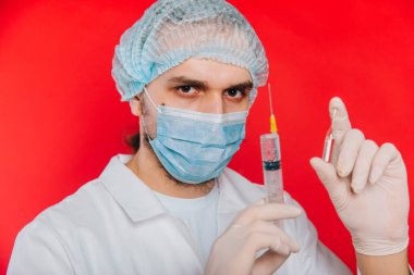 Doktor elinde şırınga ve aşılı ampul tutuyor. Kırmızı arka planda, beyaz önlüklü, tıbbi maskeli ve eldivenli genç bir adam..