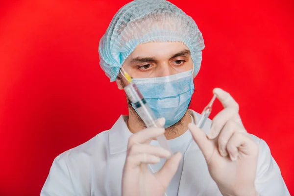 Le médecin tient une seringue et une ampoule avec un vaccin. Un jeune homme en manteau blanc, avec un masque médical et des gants sur fond rouge . — Photo