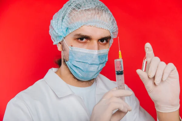 El médico sostiene una jeringa y una ampolla con una vacuna. Un joven con un abrigo blanco, con una máscara médica y guantes sobre un fondo rojo . — Foto de Stock