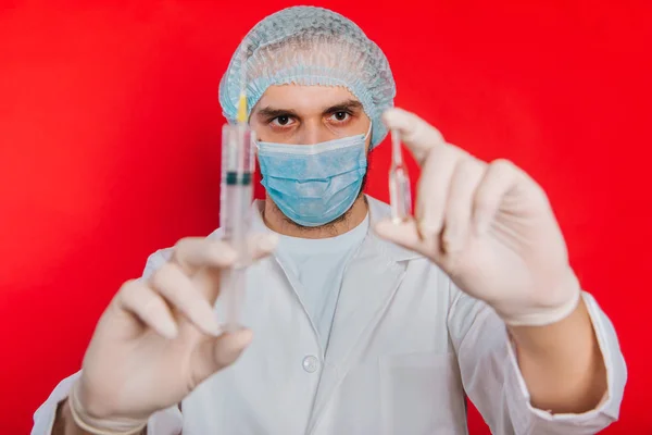 Ο γιατρός κρατάει μια σύριγγα και μια φύσιγγα με ένα εμβόλιο. Ένας νεαρός με λευκό παλτό, με ιατρική μάσκα και γάντια σε κόκκινο φόντο.. — Φωτογραφία Αρχείου