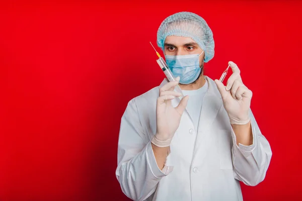 Ο γιατρός κρατάει μια σύριγγα και μια φύσιγγα με ένα εμβόλιο. Ένας νεαρός με λευκό παλτό, με ιατρική μάσκα και γάντια σε κόκκινο φόντο.. — Φωτογραφία Αρχείου