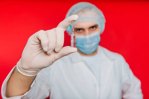 El médico sostiene en sus manos una ampolla con una vacuna. Un joven con un abrigo blanco, con una máscara médica y guantes sobre un fondo rojo. Trabajador médico . — Foto de Stock