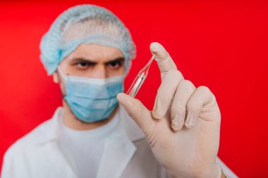 Doktor elinde bir ampul ile bir aşı tutuyordu. Beyaz önlüklü, tıbbi maskeli ve kırmızı arka planda eldivenleri olan genç bir adam. Sağlık çalışanı.