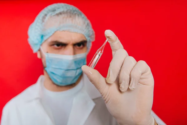 El médico sostiene en sus manos una ampolla con una vacuna. Un joven con un abrigo blanco, con una máscara médica y guantes sobre un fondo rojo. Trabajador médico . — Foto de Stock