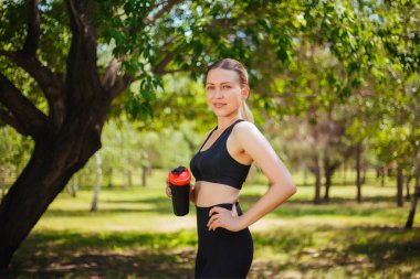 Parkta yeşil bir paspasın üzerinde elinde bir bardak suyla oturan spor kıyafetli güzel bir kız. Kulaklıkla yoga yapıyorum. Spor sırasında müzik dinler.