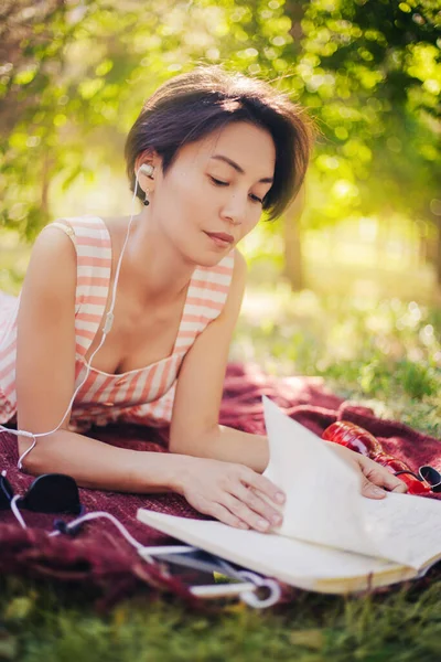 少女は草の上に横たわり、ノートに講義を読む。女性が本を読み、公園で音楽を聴く。学外教育 — ストック写真