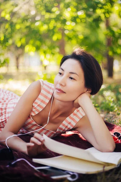 少女は草の上に横たわり、ノートに講義を読む。女性が本を読み、公園で音楽を聴く。学外教育 — ストック写真