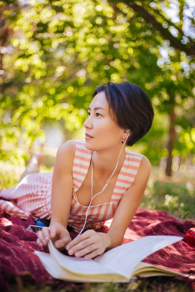 The girl lies on the grass and reads lectures in a notebook. A woman reads a book and listens to music in the park. Outdoor education