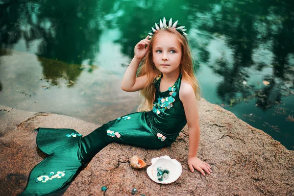 Маленькая девочка с седыми волосами с хвостом русалки и раковинами сидит на берегу. Маленькая русалочка сидит на большом камне у пруда . — стоковое фото