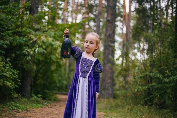 A little girl with white hair in a purple medieval dress stands with a lamp. The fairy princess got lost in the dark forest. — Stock Photo, Image