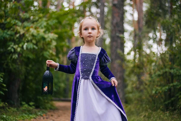 Маленькая девочка с белыми волосами в фиолетовом средневековом платье стоит с лампой. Фея принцесса заблудилась в темном лесу. . — стоковое фото