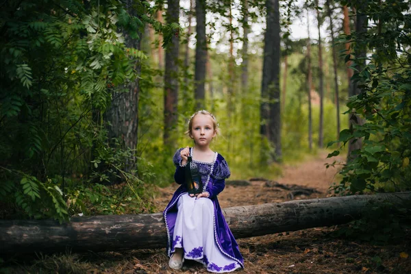 Une petite fille aux cheveux blancs dans une robe médiévale pourpre s'assoit sur un arbre avec une lampe. La princesse des fées s'est perdue dans la forêt sombre . — Photo
