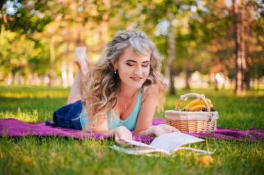 Parkta piknikte güzel bir kız kitap okuyor. Mor yatak örtüsünde dinlenen sarışın. Piknikte meyve sepeti