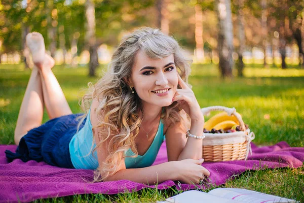 Krásná dívka na pikniku v parku čte knihu. Blondýnka na purpurovém povlečení. Koš na ovoce na pikniku — Stock fotografie