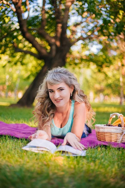 公園のピクニックの美しい少女は本を読んでいる。紫色のベッドの上にブロンドが眠っています。ピクニックのフルーツバスケット — ストック写真