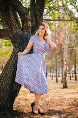 Kırmızı dudaklı ve parktaki ağaçların arka planında bezelye desenli elbisesi olan bir kız. Pin-up modeli.