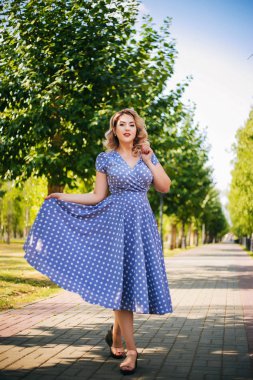 Kırmızı dudaklı ve parktaki ağaçların arka planında bezelye desenli elbisesi olan bir kız. Pin-up modeli.