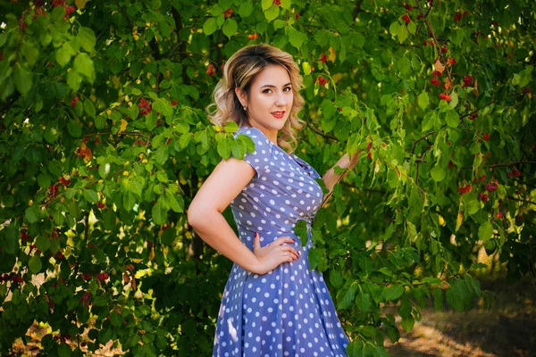 Meisje met rode lippen en een jurk in erwten op een achtergrond van bomen in het park. Pin-up model op een achtergrond van rode appels. — Stockfoto