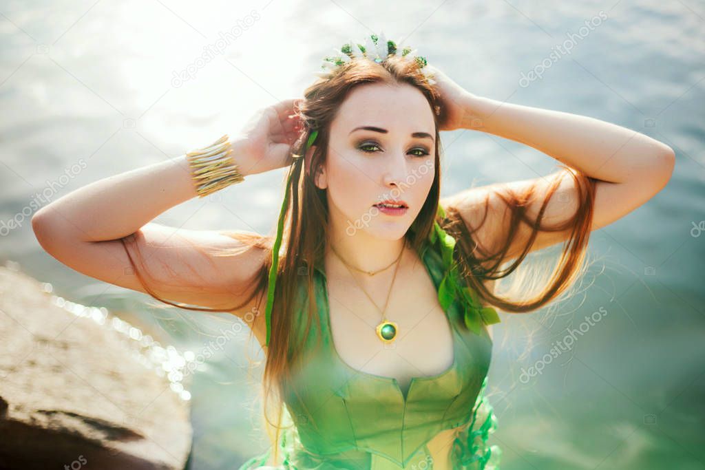 A girl with seaweed in her hair and in a green dress is standing near a pond. Model with a rim with shells. Mermaid