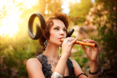 A girl in the image of a faun plays the wooden flute. Model with horns on his head and with furry pants in the woods clipart