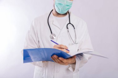 Tıbbi maskeli beyaz önlüklü doktor elinde arka planda kalem olan mavi bir dosya tutuyor..