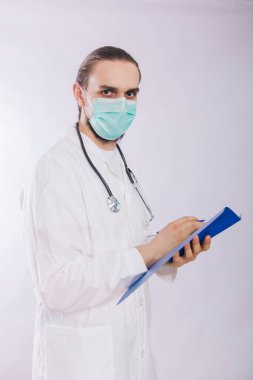 Tıbbi maskeli beyaz önlüklü doktor elinde arka planda kalem olan mavi bir dosya tutuyor..