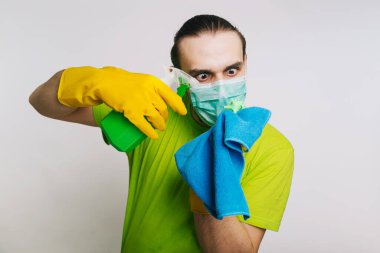 Yeşil tişörtlü ve tıbbi maskeli bir adam elinde antiseptik ve beyaz arka planda toz bezi olan bir bulletzer tutuyor. Bir adam koronavirüsü öldürür..