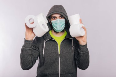 Gri kazaklı ve tuvalet kağıdı ruloları beyaz arka planda duran bir adam. Koronavirüs karantinası sırasındaki heyecan. Tıbbi maskeli bir adamda Rotavirüsler.